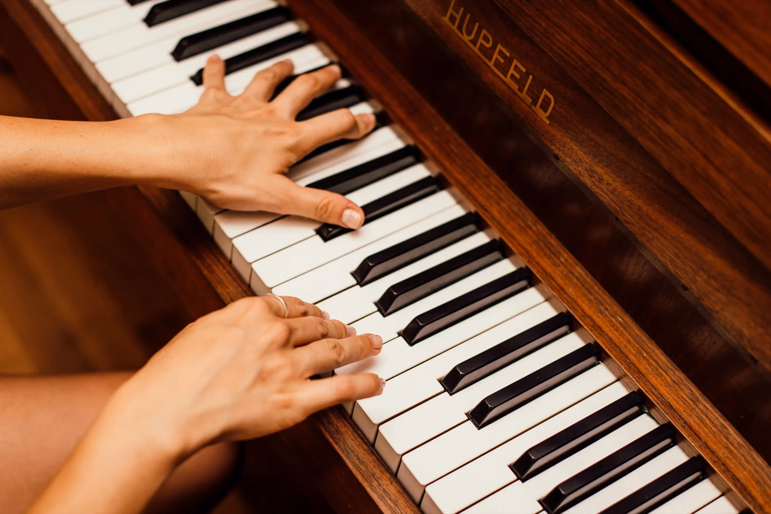 A close-up view of hands playing on an elegant wooden piano with "Hupfeld" inscribed on its side.
