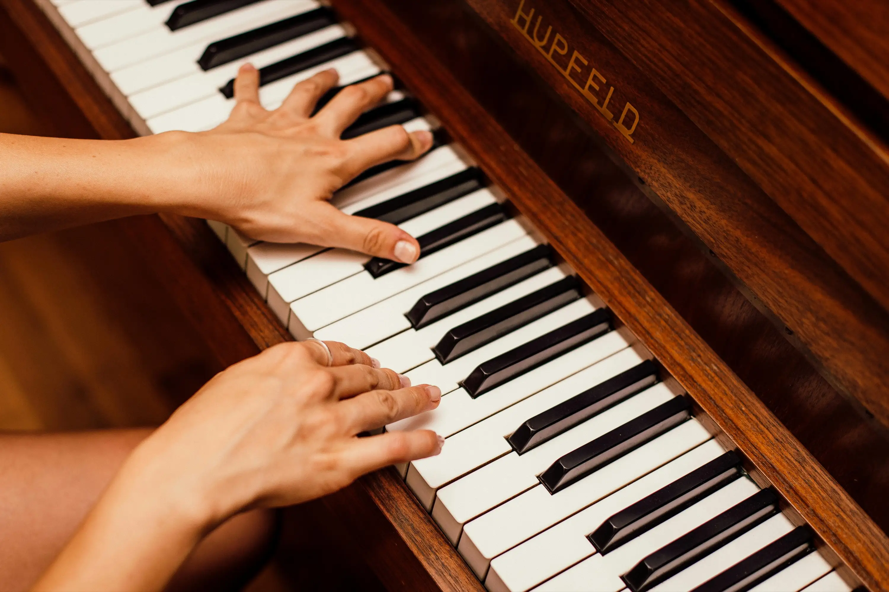 A close-up view of hands playing on an elegant wooden piano with "Hupfeld" inscribed on its side.