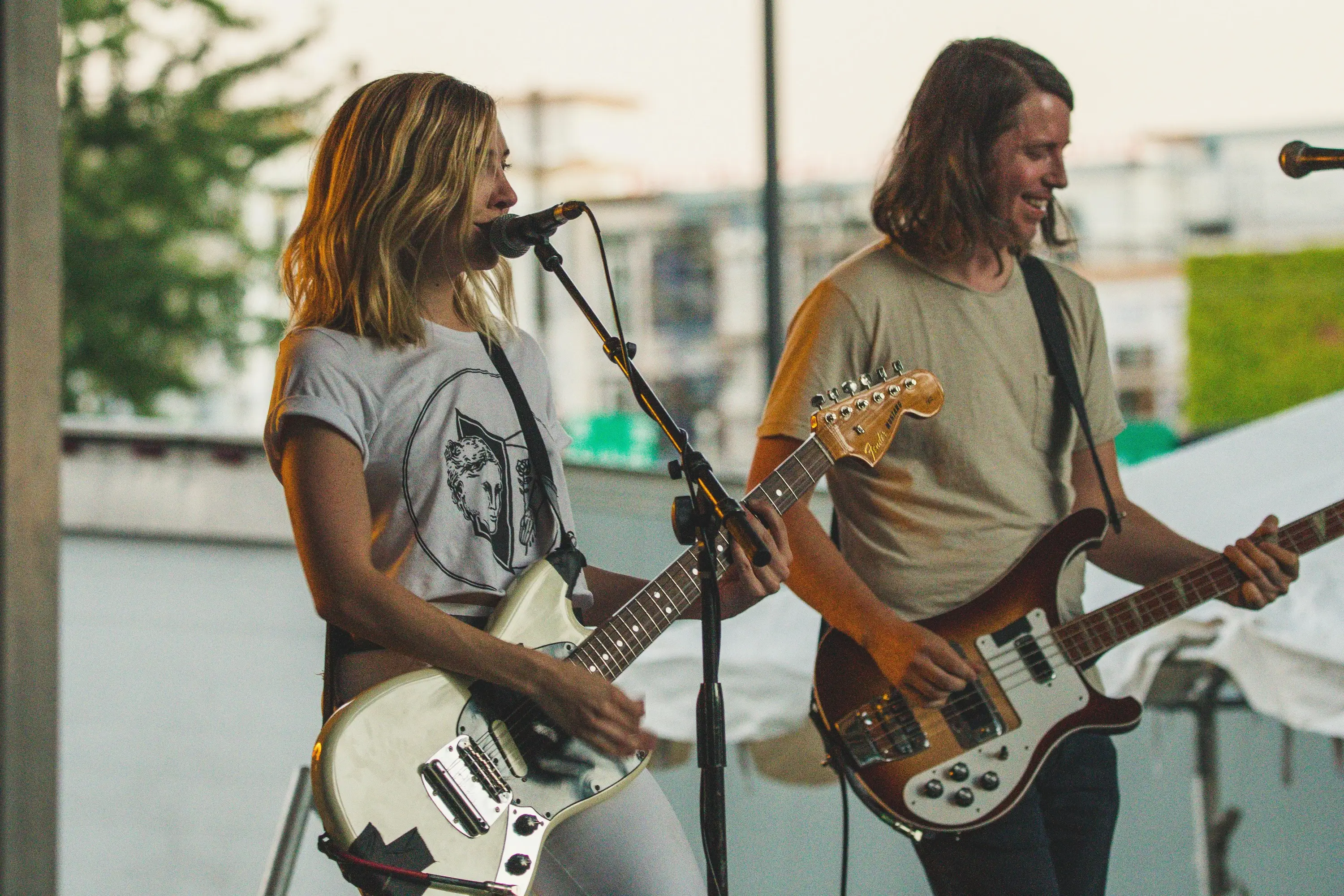 Two musicians are playing guitars and smiling while performing outdoors, with a microphone in front of them.