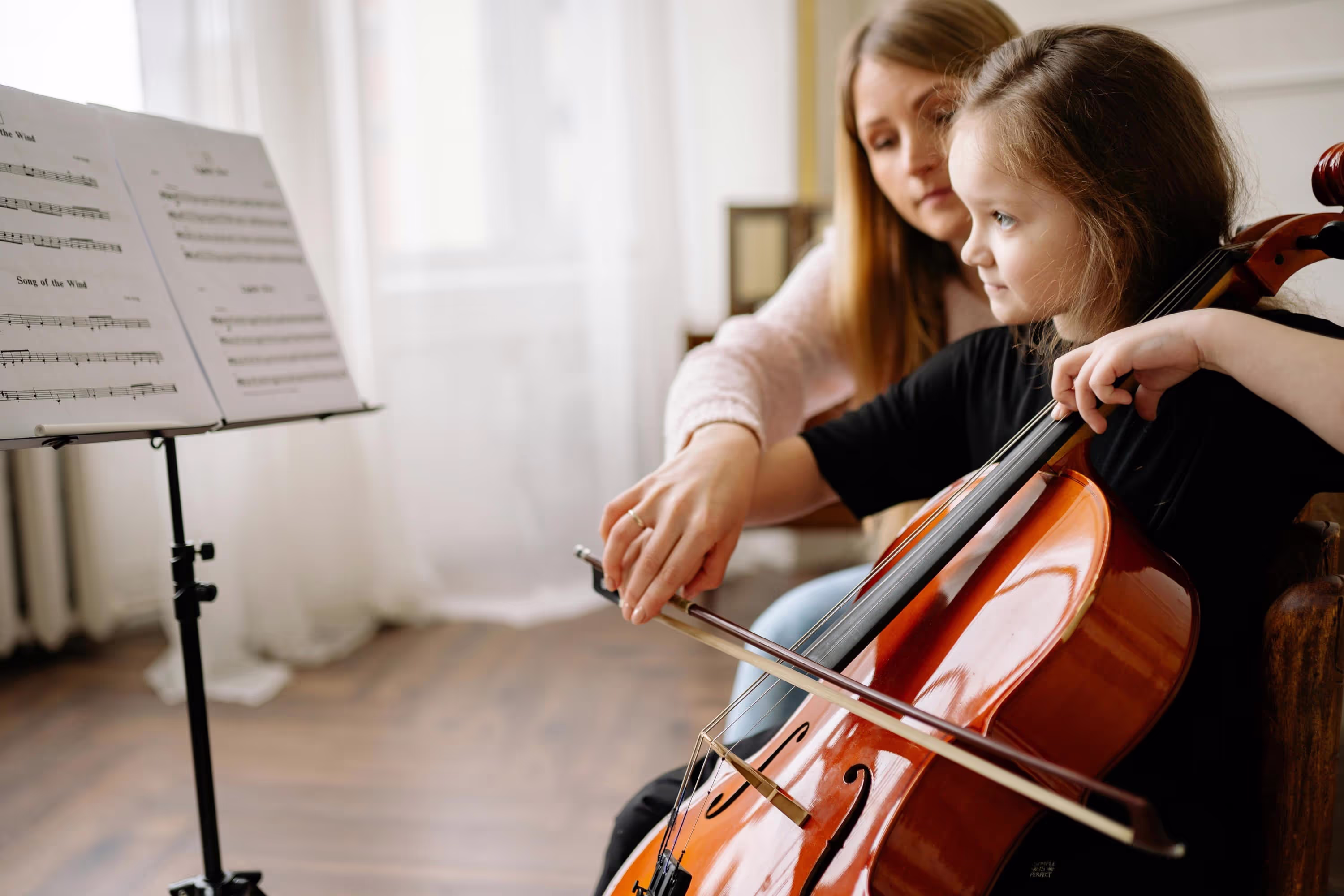 A woman teaches a young girl to play the cello in a cozy, well-lit room with sheet music nearby.