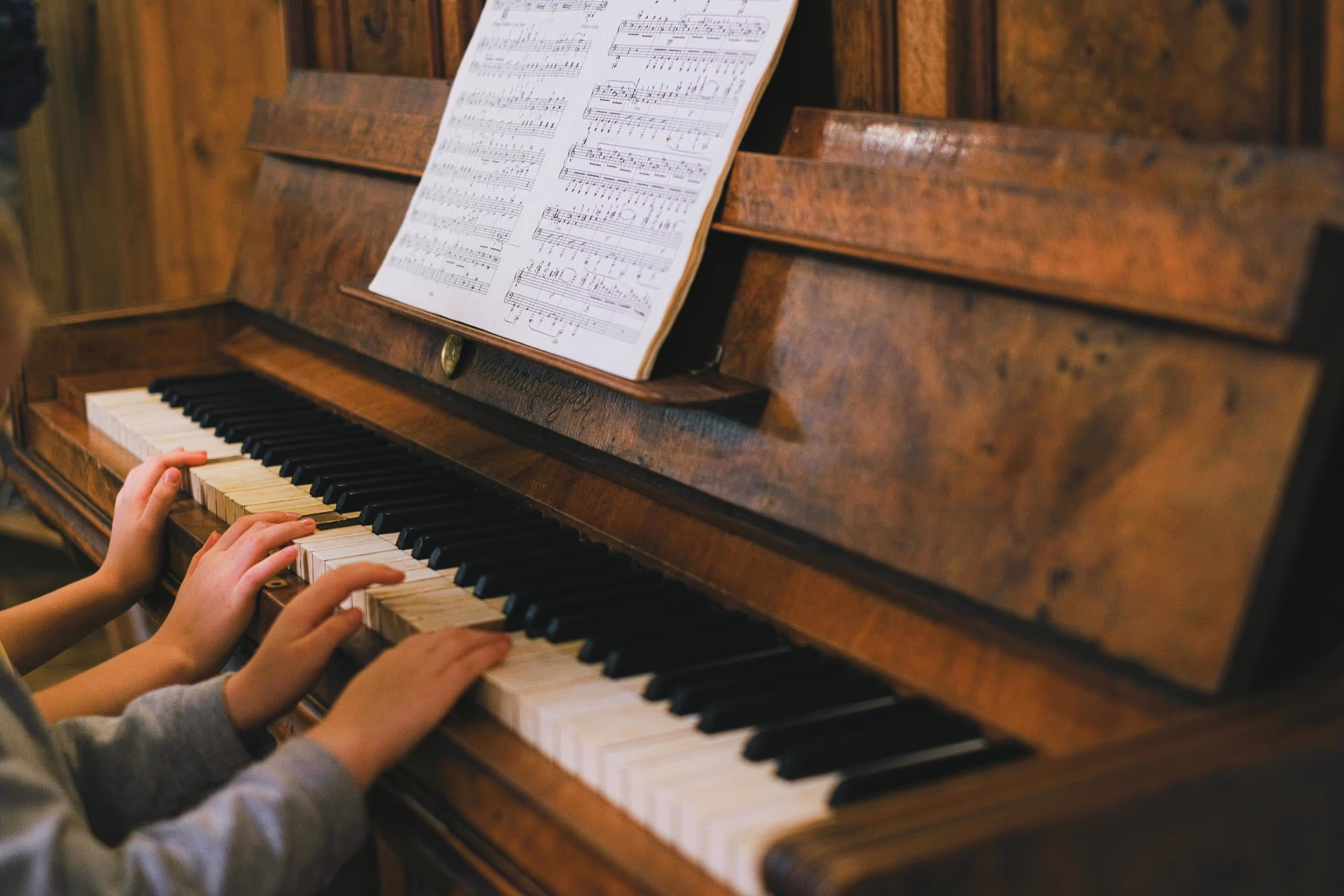 A child's hands gently press keys on an old, wooden piano with sheet music resting above it.