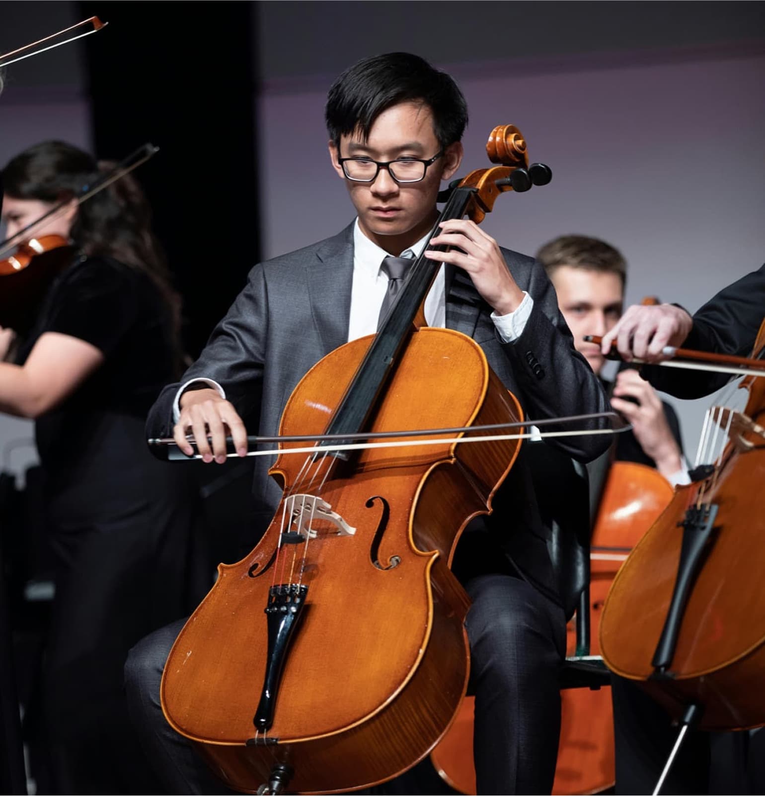 A musician in formal attire plays the cello with other string players in the background during an orchestral performance.