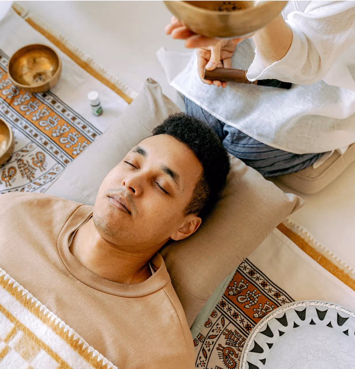 A person is receiving a soothing sound therapy session with singing bowls, lying on a cushioned surface in a tranquil setting.