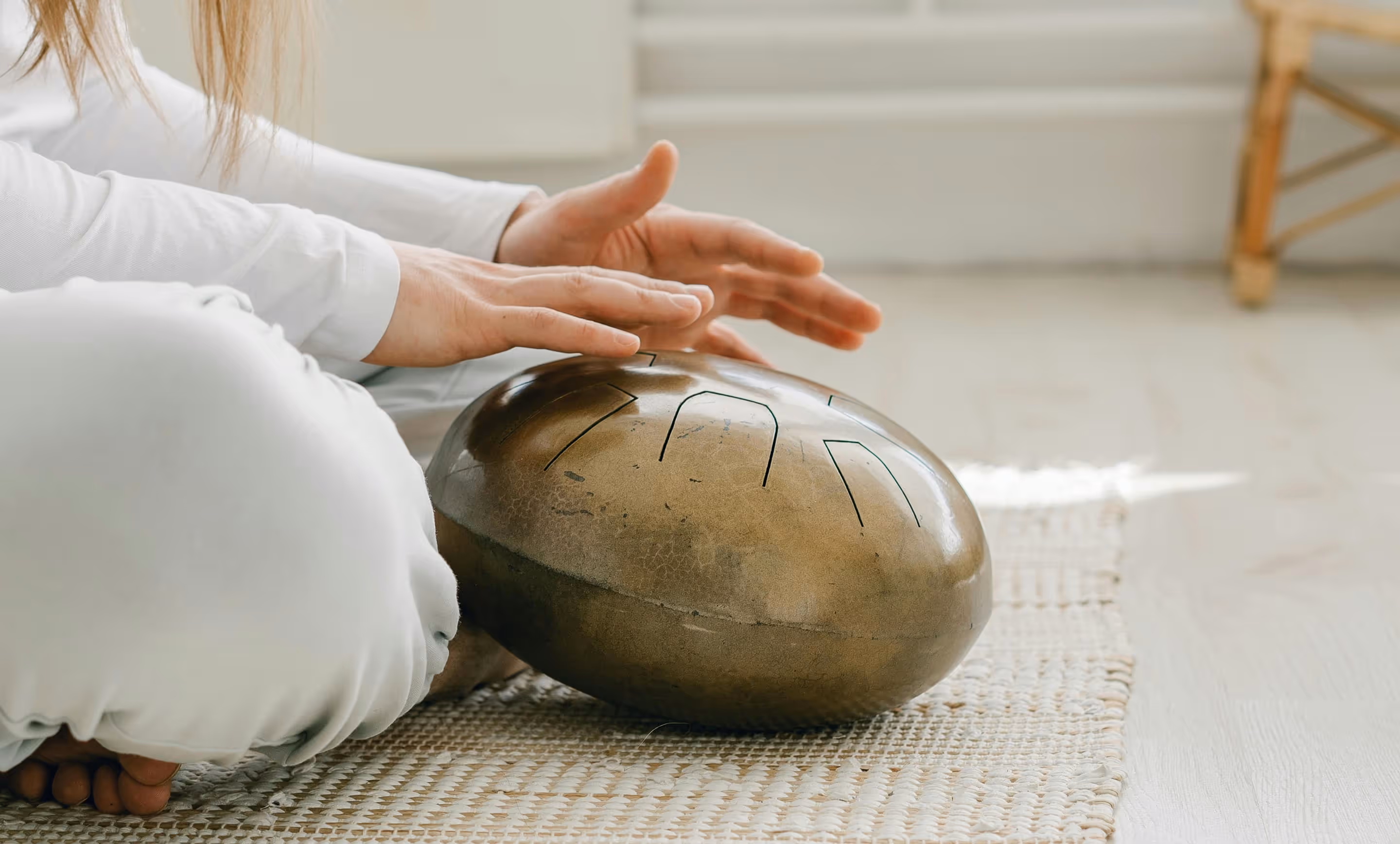 A person in white attire sits cross-legged on a woven mat, gently resting their hands atop a bronze tongue drum.