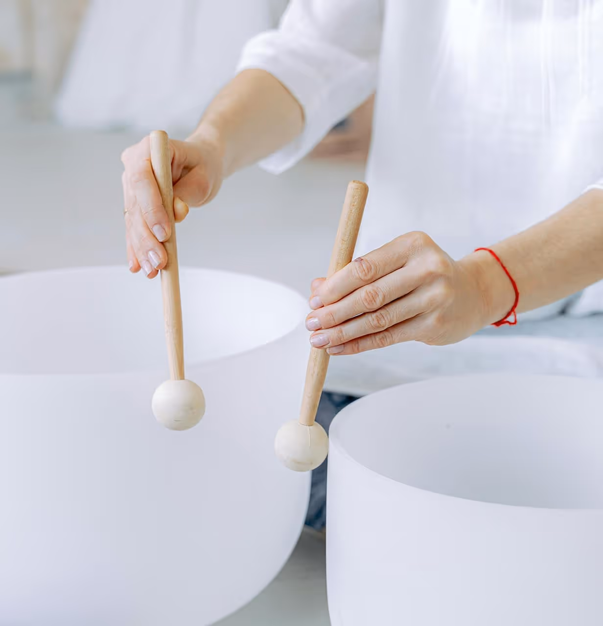 A person holding wooden mallets poised above two white crystal bowls, ready to play or perform with them.